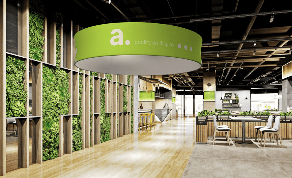 Modern indoor space with green walls, wooden flooring, and a large circular ceiling light featuring "a. quality on display." Bar area with seating is visible on the right.